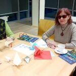 Kobieta siedzi przy stole zastawionym papierem kolorowym i wyrobami origami podczas warsztatów.