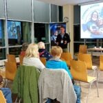 Uczestnicy spotkania "Cyfrowy Senior" słuchają prelekcji eksperta stojącego przed ekranem projekcyjnym w sali bibliotecznej.