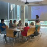 Grupa seniorów uczestniczy w warsztatach fotograficznych, słuchając prezentacji przy projekcji na ekranie.