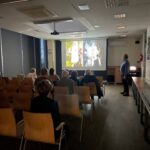 Uczestnicy wykładu siedzą w sali z projektorem wyświetlającym prezentację dotyczącą symboliki koronacji.