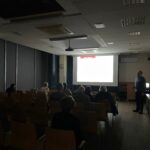 W sali wykładowej zgromadzeni słuchacze obserwują prezentację wyświetlaną na ekranie.