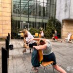 Uczestnicy zajęć jogi wykonują ćwiczenia siedząc na krzesłach przed budynkiem Miejskiej Biblioteki Publicznej w Chrzanowie.