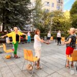 Uczestnicy zajęć jogi wykonują ćwiczenia przy wykorzystaniu krzeseł na zewnątrz biblioteki.