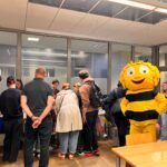 Uczestnicy wydarzenia otaczają maskotkę pszczoły stojącą na stole wewnątrz biblioteki.