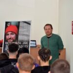 Mężczyzna w zielonej koszuli przemawia do publiczności, a za nim widoczny jest plakat z hasłem „Betonizacja jak szczury”.