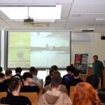 Uczniowie słuchają prezentacji wyświetlanej na ekranie projekcyjnym w sali wykładowej.