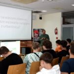 Mężczyzna wygłasza wykład przed grupą uczniów siedzących w sali szkolnej.