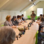 Uczestnicy warsztatów siedzą przy długim stole, na którym znajdują się materiały do tworzenia biżuterii.