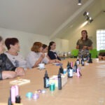 Uczestniczki warsztatów z żywicy epoksydowej słuchają wykładu prowadzącej, siedząc przy długim stole zastawionym materiałami do pracy.