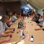 Uczestniczki warsztatów rękodzielniczych siedzą przy długim stole, na którym znajdują się butelki z żywicą i narzędzia do tworzenia biżuterii.