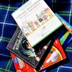Na tle piknikowej tkaniny leżą kolorowe książki dla dzieci o tematyce przyrodniczej.