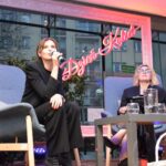 Dwie kobiety siedzą na krzesłach podczas programu telewizyjnego, jedna trzyma w ręku filiżankę.