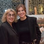 Dwie kobiety w czarnych marynarkach pozują do zdjęcia na tle wystawy sztuki.