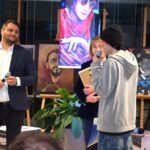 Mężczyźni prowadzą rozmowę przy mikrofonie w towarzystwie wystawionych obrazów, wśród których widoczne są portrety inspirowane The Beatles.