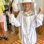 Dziecko uśmiecha się, ubrane w strój pszczelarski podczas zajęć z lokalnym pszczelarzem na Kulturalnych Wakacjach.