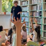 Nauczycielka podczas zajęć z dziećmi w bibliotece, dzieci podnoszą ręce.