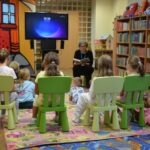 Kobieta czyta dzieciom książkę siedzącym na zielonych krzesłach podczas spotkania w bibliotece.