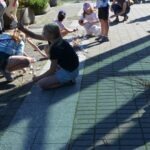 Dzieci siedzą na podłożu z gumowych kostek podczas zajęć edukacyjnych na świeżym powietrzu.
