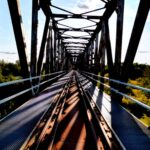 Widok z perspektywy na szyny kolejowe biegnące przez metalowy most nad rzeką w słoneczny dzień.