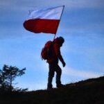 Mężczyzna w plecaku niesie flagę Polski na wzgórzu oświetlonym zachodzącym słońcem.