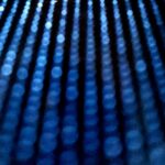 Abstrakcyjne zdjęcie w odcieniach błękitu przedstawiające pionowe linie świetlne, przypominające strumienie wody lub światła lasera.