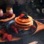Stos pancakes z truskawkami i jagodami na talerzu, otoczony kwiatami i liśćmi.