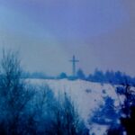 Zasneżony wzgórze z krzyżem na szczycie, otoczone drzewami w pochmurny dzień.