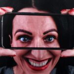 Kobieta uśmiecha się szeroko i trzyma telefon przed twarzą, powiększając swoje oczy.
