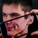 Mężczyzna trzyma smartfon przed twarzą, na ekranie widoczne jego odbicie.