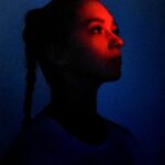 Portret kobiety z ciemną skórą i splecionymi włosami oświetlony czerwonym i niebieskim światłem.