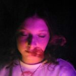 Portret młodej kobiety w ciemnym otoczeniu oświetlonym fioletowym światłem, z widoczną biżuterią na szyi.