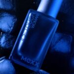Butelka perfum Mexx Ice Touch Woman leży na ciemnych kamieniach i muszelkach, oświetlona niebieskawym światłem.