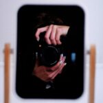 Odbicie osoby trzymającej aparat fotograficzny w lustrze oprawionym w drewnianą ramę.