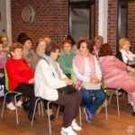 Grupa osób siedzi na krzesłach podczas wydarzenia kulturalnego w bibliotece.