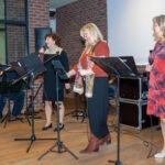 Trzy kobiety z zespołem stoją za mikrofonami, trzymając kartki z tekstem piosenki.