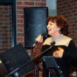 Wokalistka zespołu trzyma mikrofon i gestykuluje podczas występu na scenie.