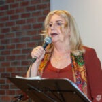 Wokalistka zespołu trzyma mikrofon i mówi przy mównicy podczas koncertu w bibliotece.