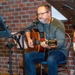 Muzyk siedzący na krześle gra na gitarze akustycznej, śpiewając do mikrofonu przed publicznością.