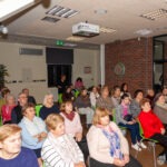 Publiczność siedzi na krzesłach, skupiona na projekcji wyświetlanej na ekranie podczas koncertu w bibliotece.