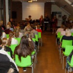 Publiczność siedzi na krzesłach w sali biblioteki, słuchając występu zespołu podczas koncertu.