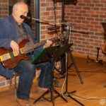 Muzyk siedzi na krześle, gra na gitarze elektrycznej i śpiewa do mikrofonu przed straganem z nutami.