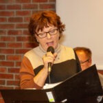 Kobieta z mikrofonem przemawia przed publicznością przy podwyższonym pulpicie.