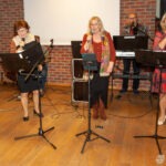 Na scenie biblioteki występują członkowie zespołu z mikrofonami i nutami, akompaniowani pianistą.