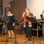 Dwie kobiety z mikrofonami stoją za stoiskami nutowymi podczas występu zespołu w bibliotece.