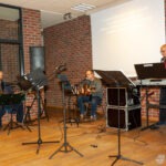 Na scenie biblioteki znajdują się członkowie zespołu z instrumentami i nutami na stojakach.