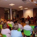 Publiczność siedzi na krzesłach, skierowana w stronę sceny, gdzie zespół prezentuje piosenki z wyświetlanymi tekstami.