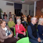 Publiczność siedzi na krzesłach, słuchając koncertu w bibliotece.