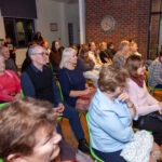 Publiczność siedzi na krzesłach, słuchając koncertu zespołu w bibliotece.