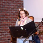 Wokalistka zespołu z mikrofonem stoi za pulpitem, prezentując tekst piosenki publiczności.