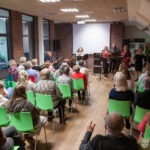 Publiczność siedzi na krzesłach, słuchając występu zespołu podczas koncertu w bibliotece.